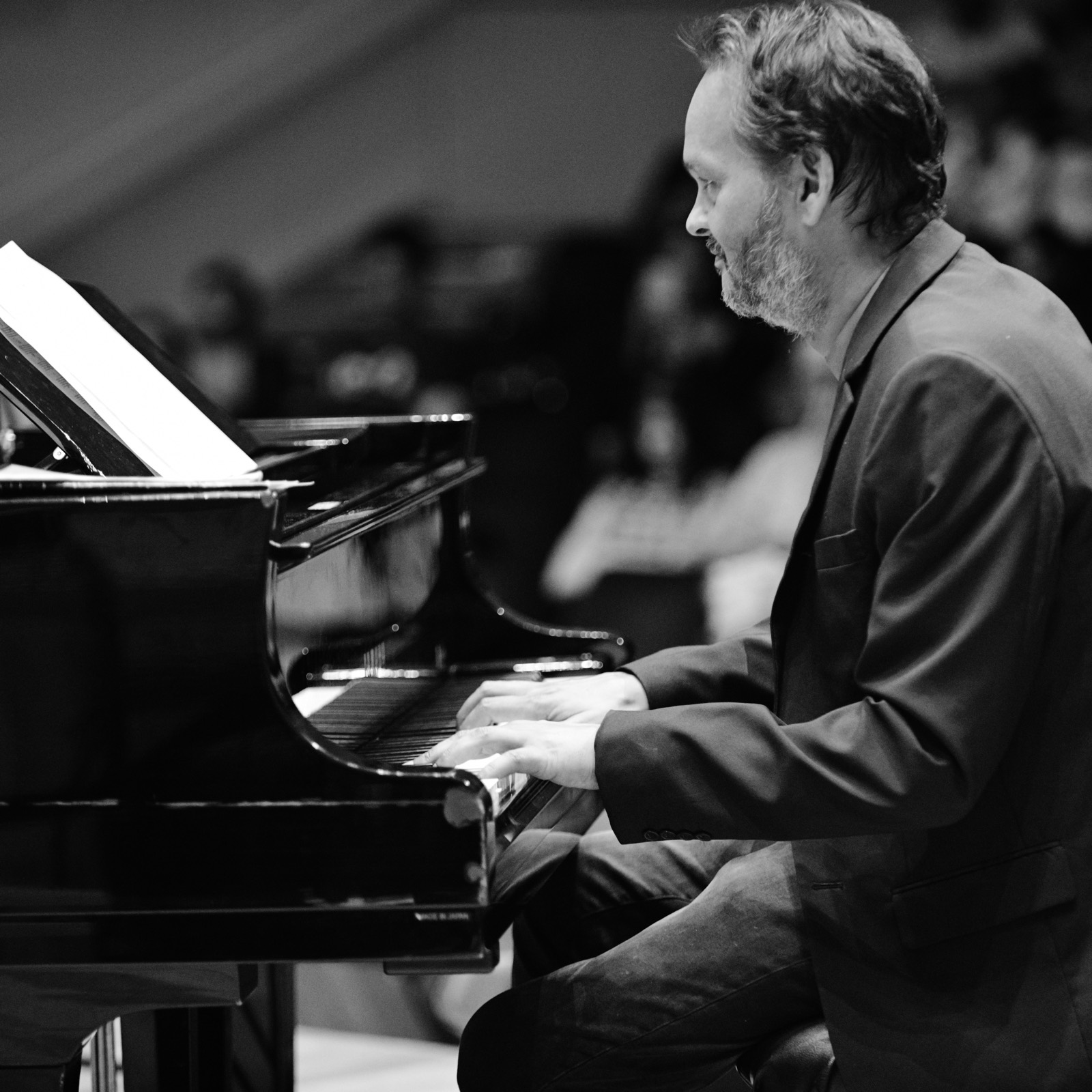 Ramon Alexander playing a grand piano in performance — a black-and-white profile with an audience behind him.