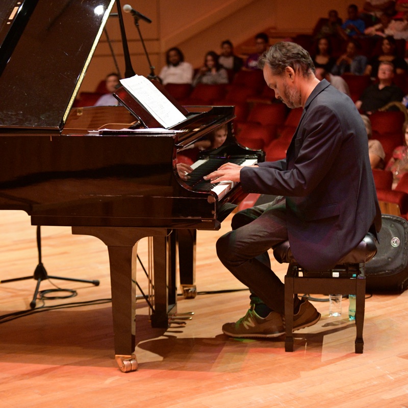 Ramon Alexander playing a grand piano on a concert-hall stage with the audience seated behind him.