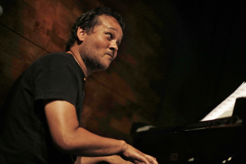 Ramon Alexander leaning into the keyboard of a grand piano, lit from above, during his residency at No Black Tie, Kuala Lumpur.
