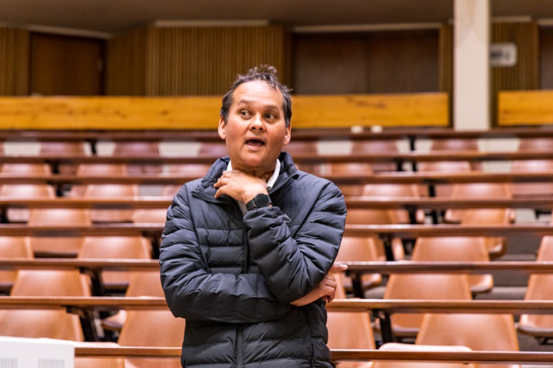 Ramon Alexander speaking and gesturing at the front of a tiered university lecture hall lined with empty wooden benches.