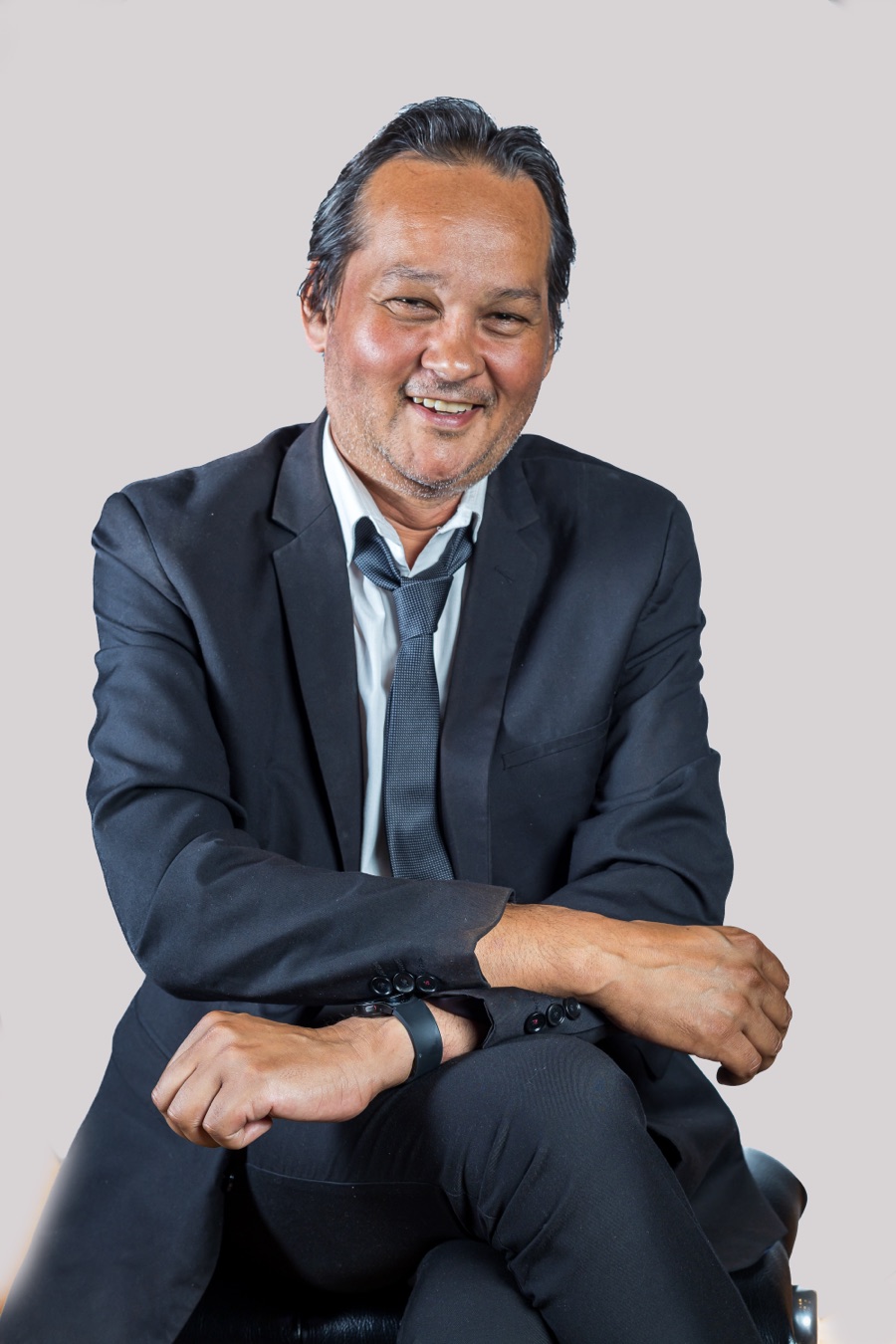Studio portrait of Ramon Alexander, seated in a dark suit with his arms resting on his knee, smiling to camera.